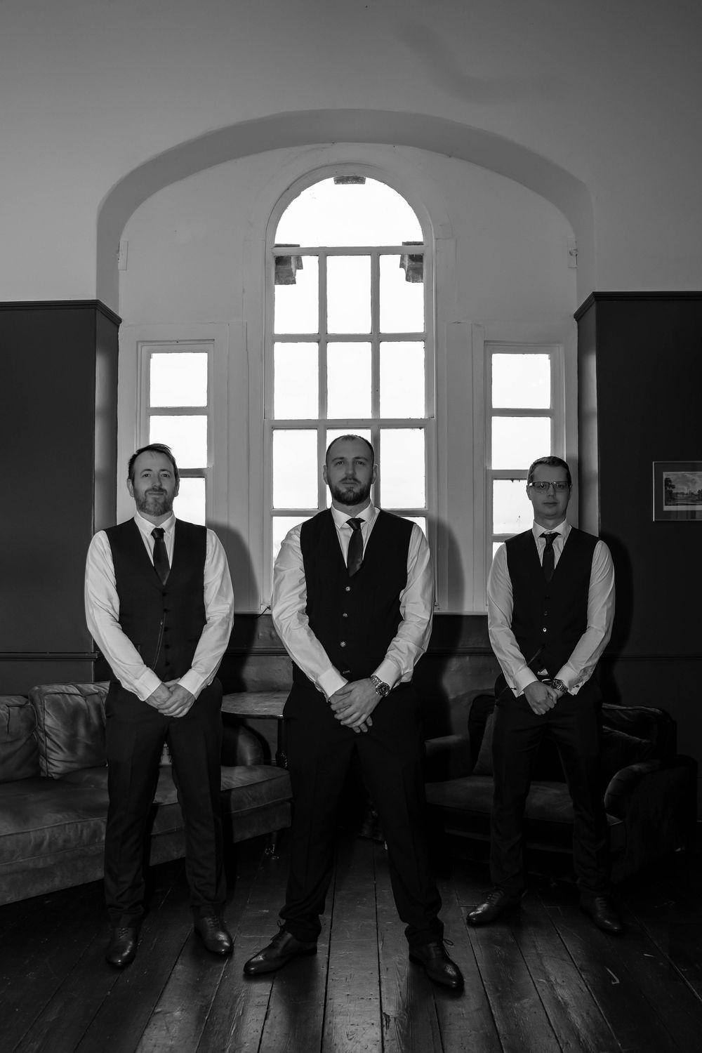 Three groomsmen in matching vests stand formally beneath an arched window.