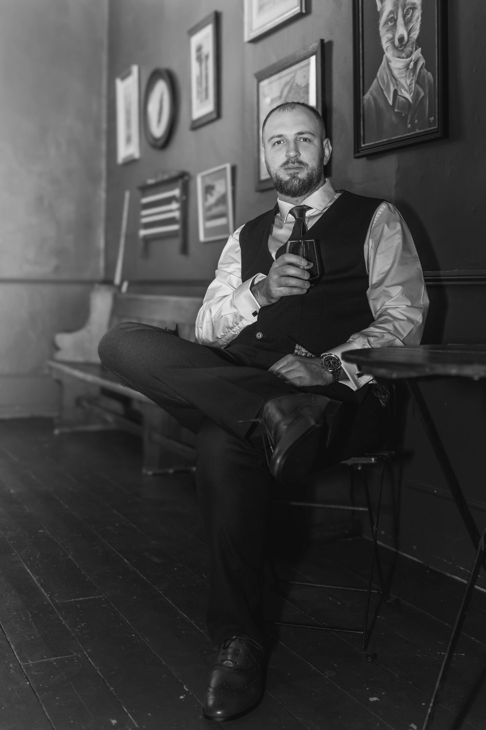 A person in formal attire sits with a drink in a vintage-style room with framed artwork.