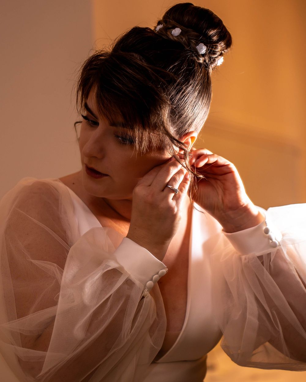 Someone adjusts their earrings while wearing a sheer white top in warm lighting.