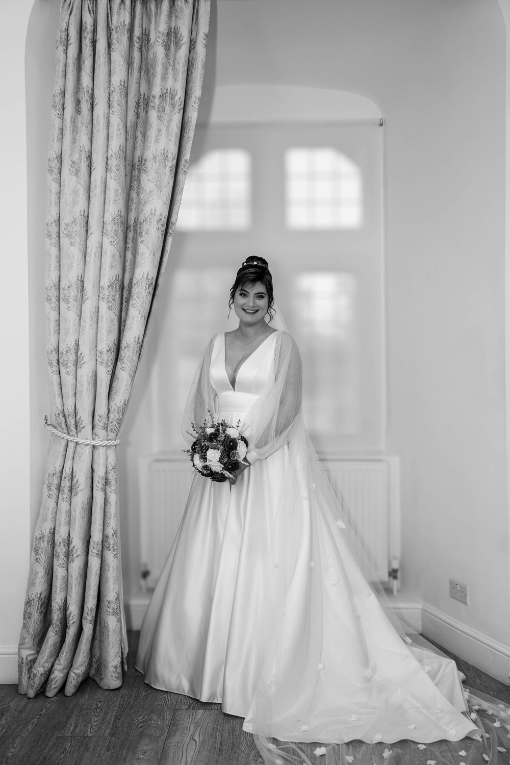 A classic bridal portrait showing a long white wedding gown next to an elegant curtained window.