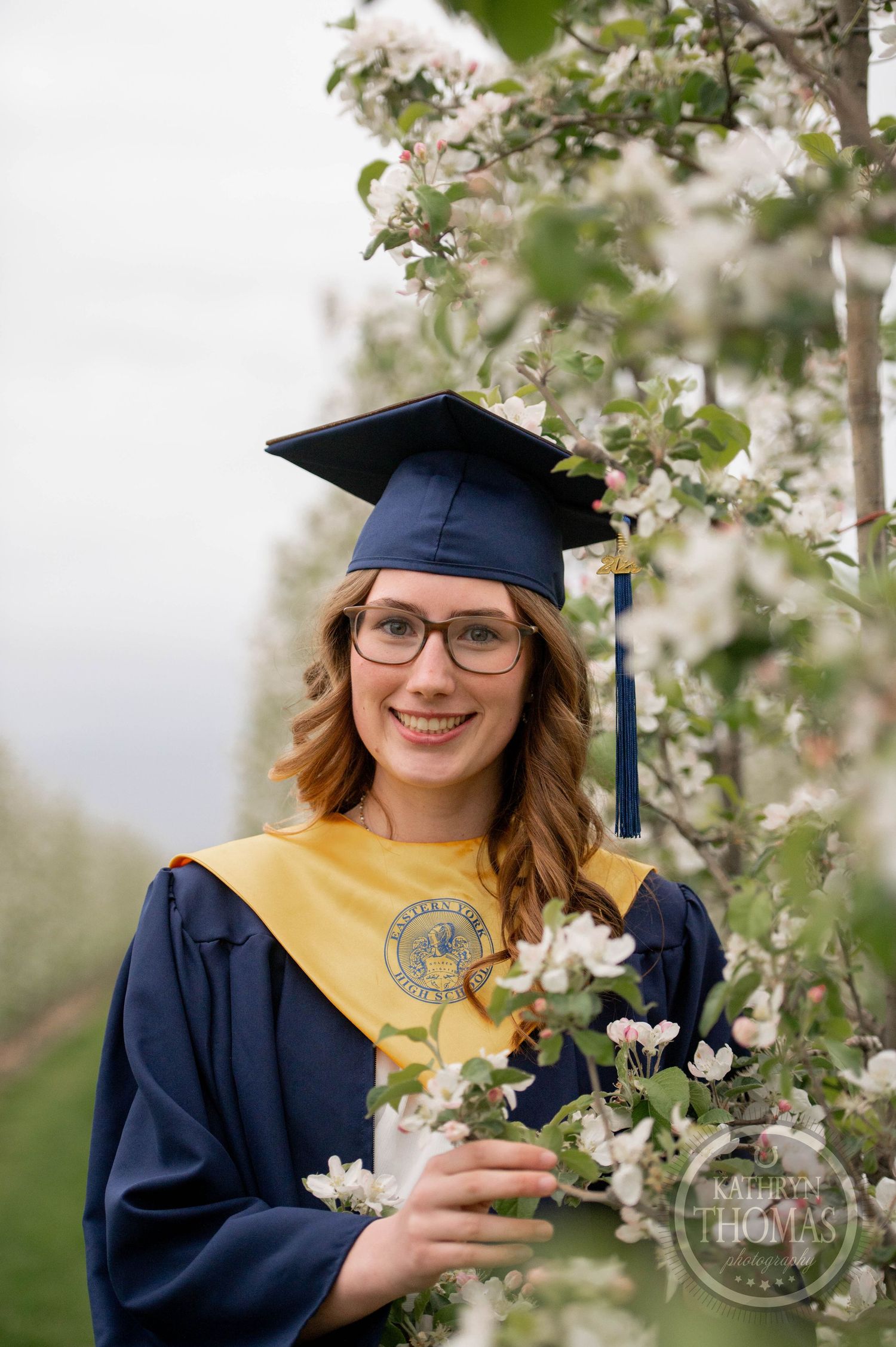 Graduate in navy cap and gown with gold stole poses next to blooming white apple blossoms in spring outdoor setting.