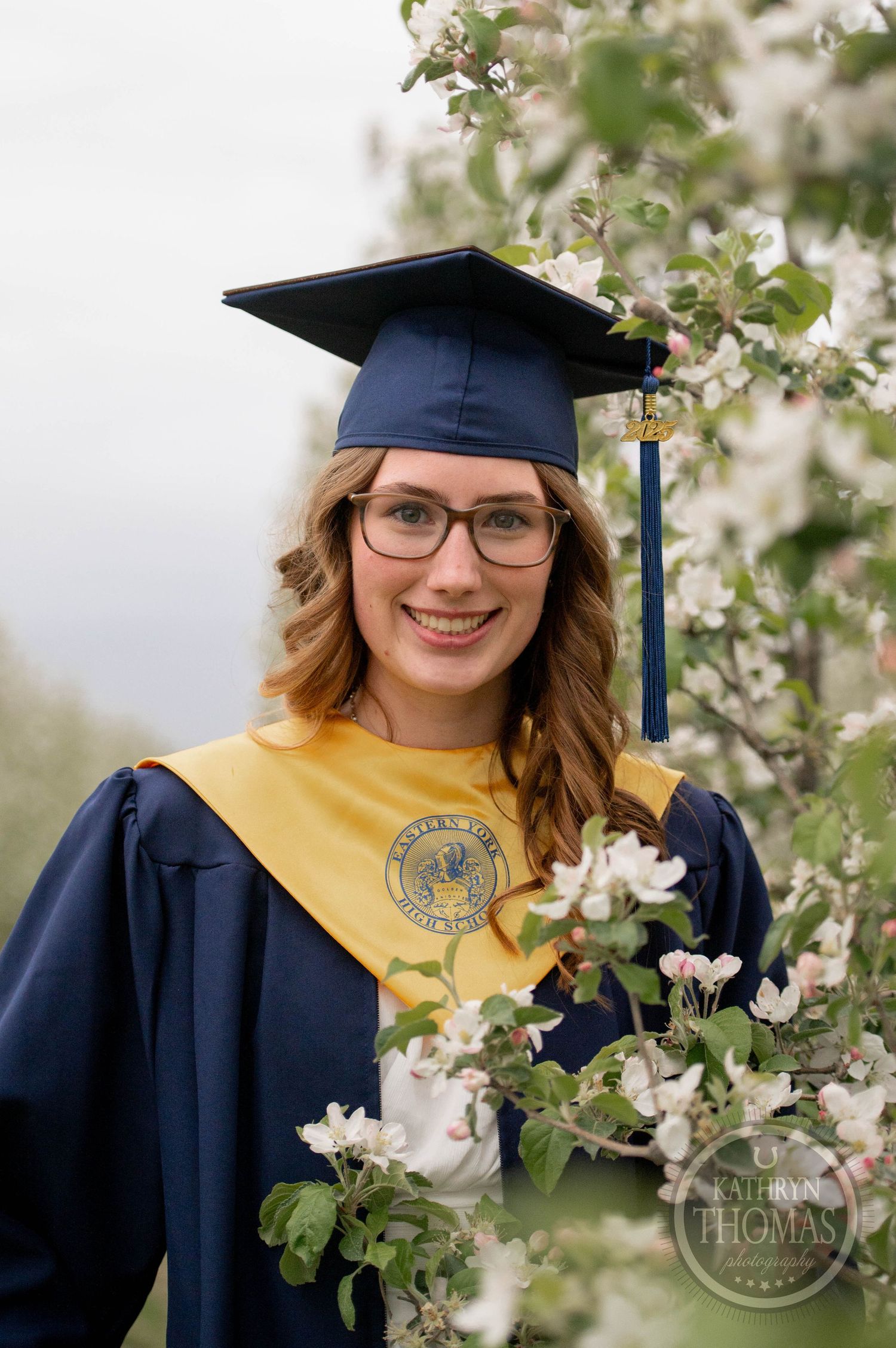 Graduate in navy cap and gown with gold honors stole poses among spring blossoms beneath flowering tree.