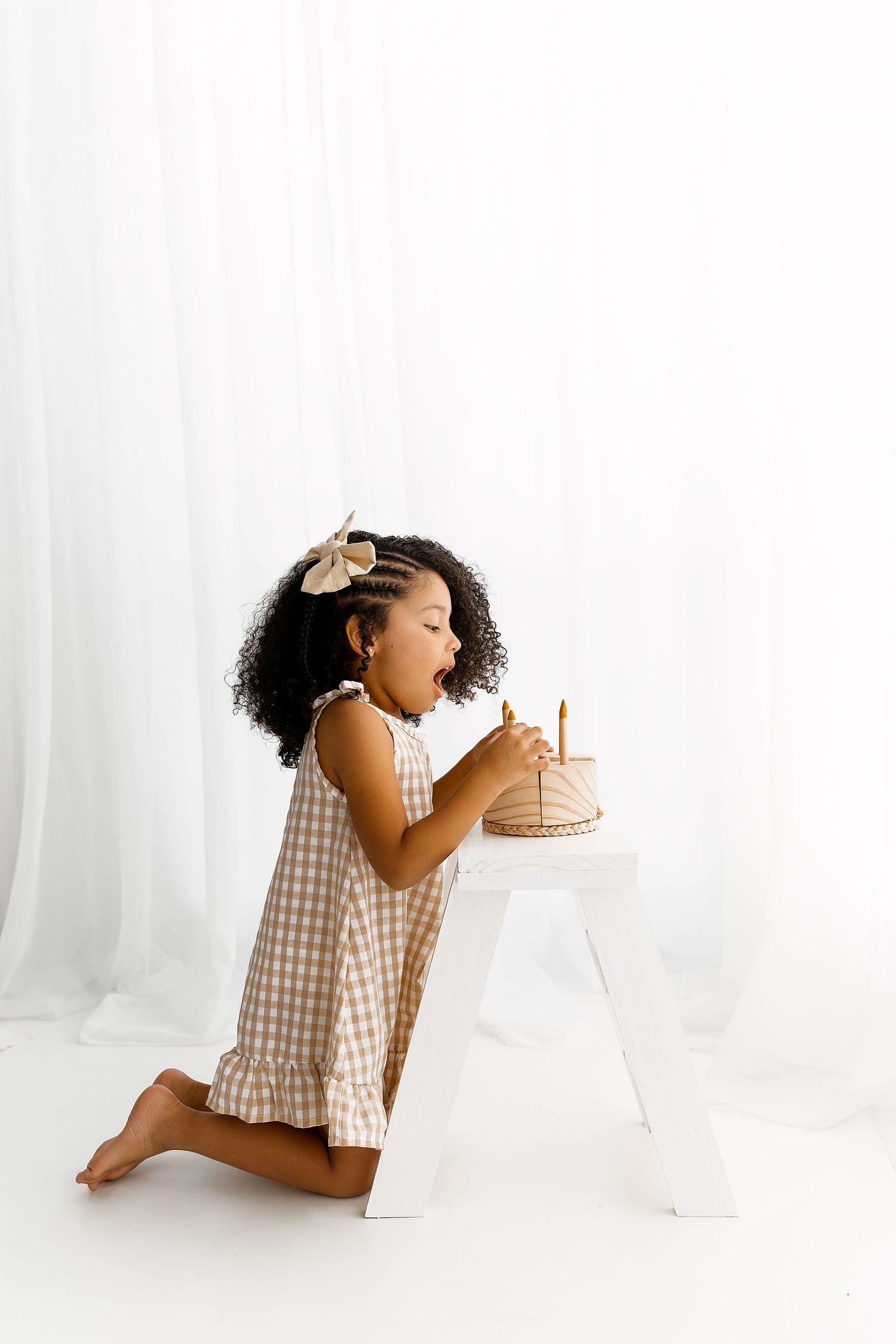 Young child in gingham dress kneels beside white letter A decoration in minimalist white studio setting.