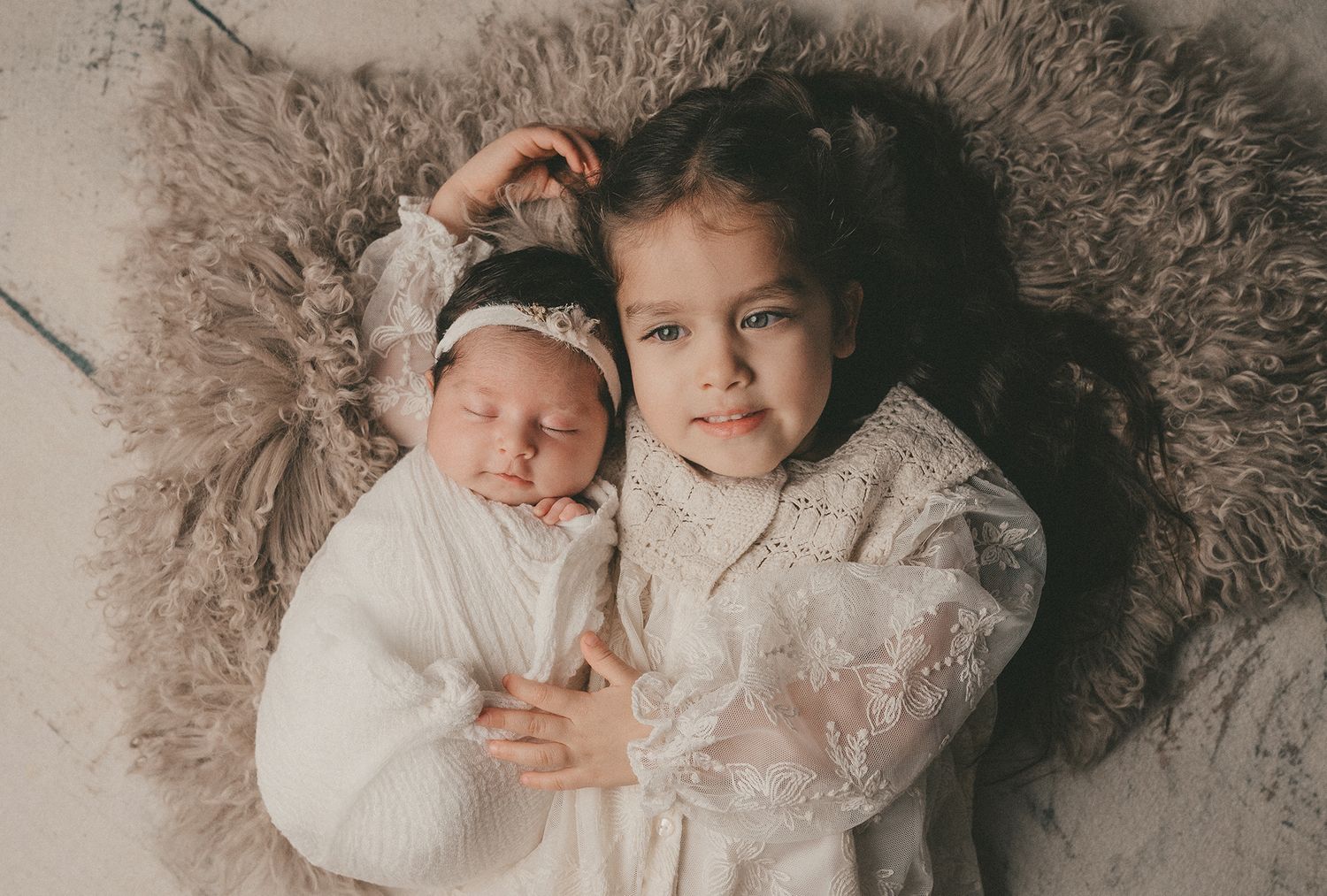 Two children in cozy white sweaters laying on plush gray furry blanket.