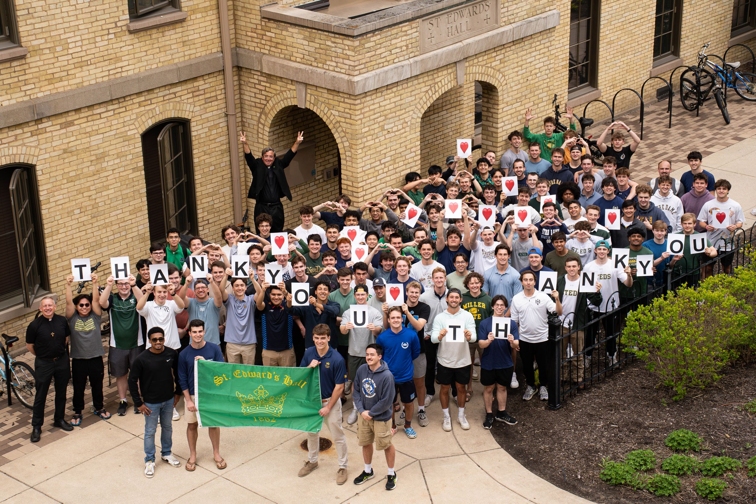 A large group of people in black and white shirts stand gathered on stone steps outside a brick building with a green banner.