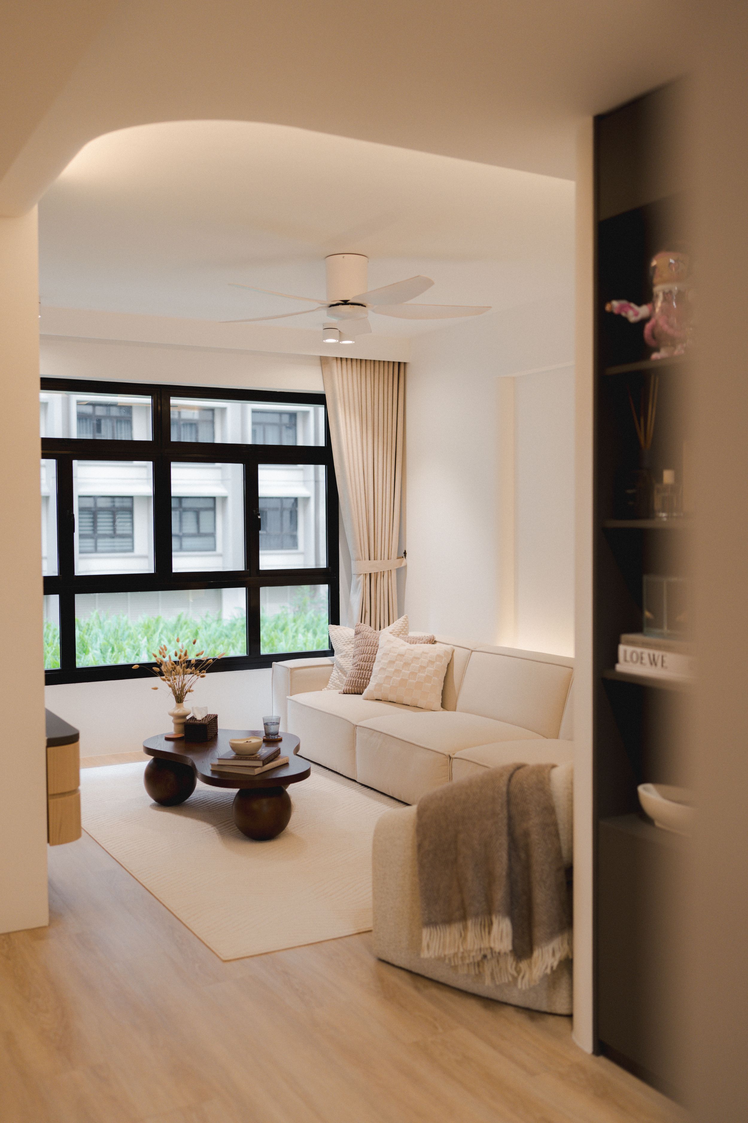 Modern minimalist living room with white sofa, wooden floors, large windows and built-in shelves in warm neutral tones.