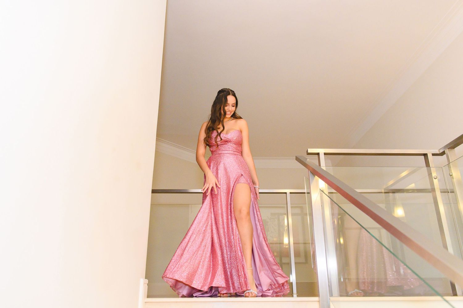 A person in a flowing pink satin evening gown with a high slit poses elegantly on a modern staircase.