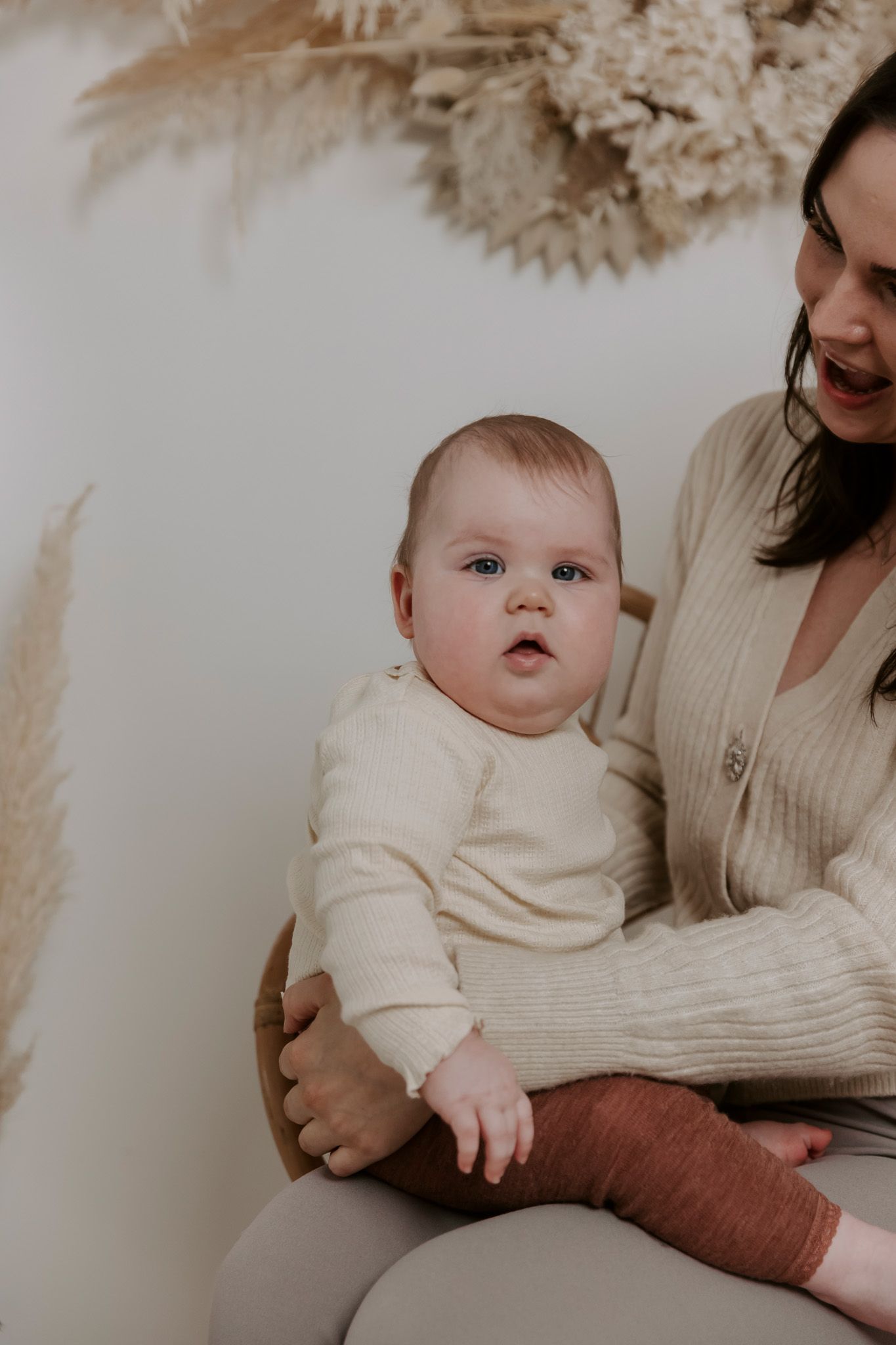 Mor og baby i neutrale toner sidder foran en hvid væg med pampasgræs som dekoration. Børnefotograf Fyn