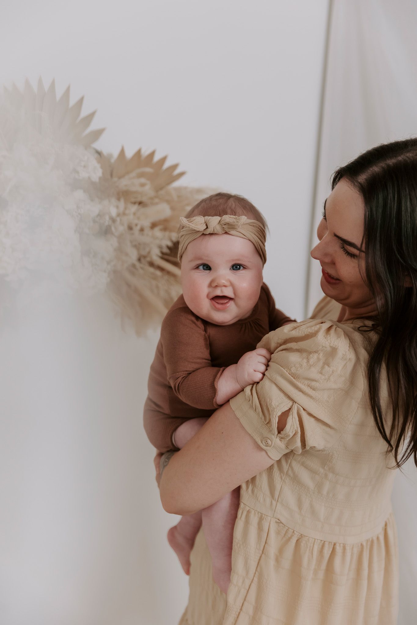 Mor og baby deler et ømt øjeblik foran en neutral baggrund med palmebladsdekoration. Børnefotograf Fyn