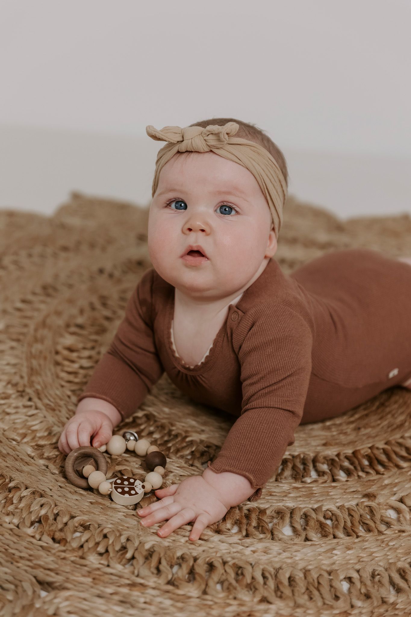 En baby i brunt outfit ligger på et tekstureret tæppe med trælegetøj. Børnefotograf Fyn