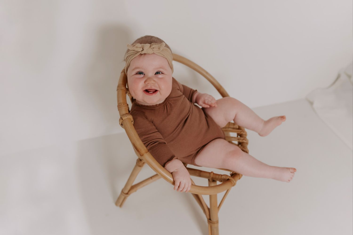 En baby iført brunt outfit hviler på rattanmøbler foran en hvid væg. Børnefotograf Fyn
