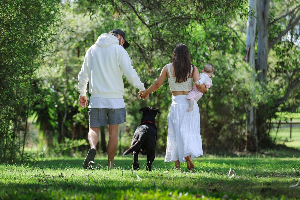 A couple walks their black dog together on a sunny day in a lush green park setting.