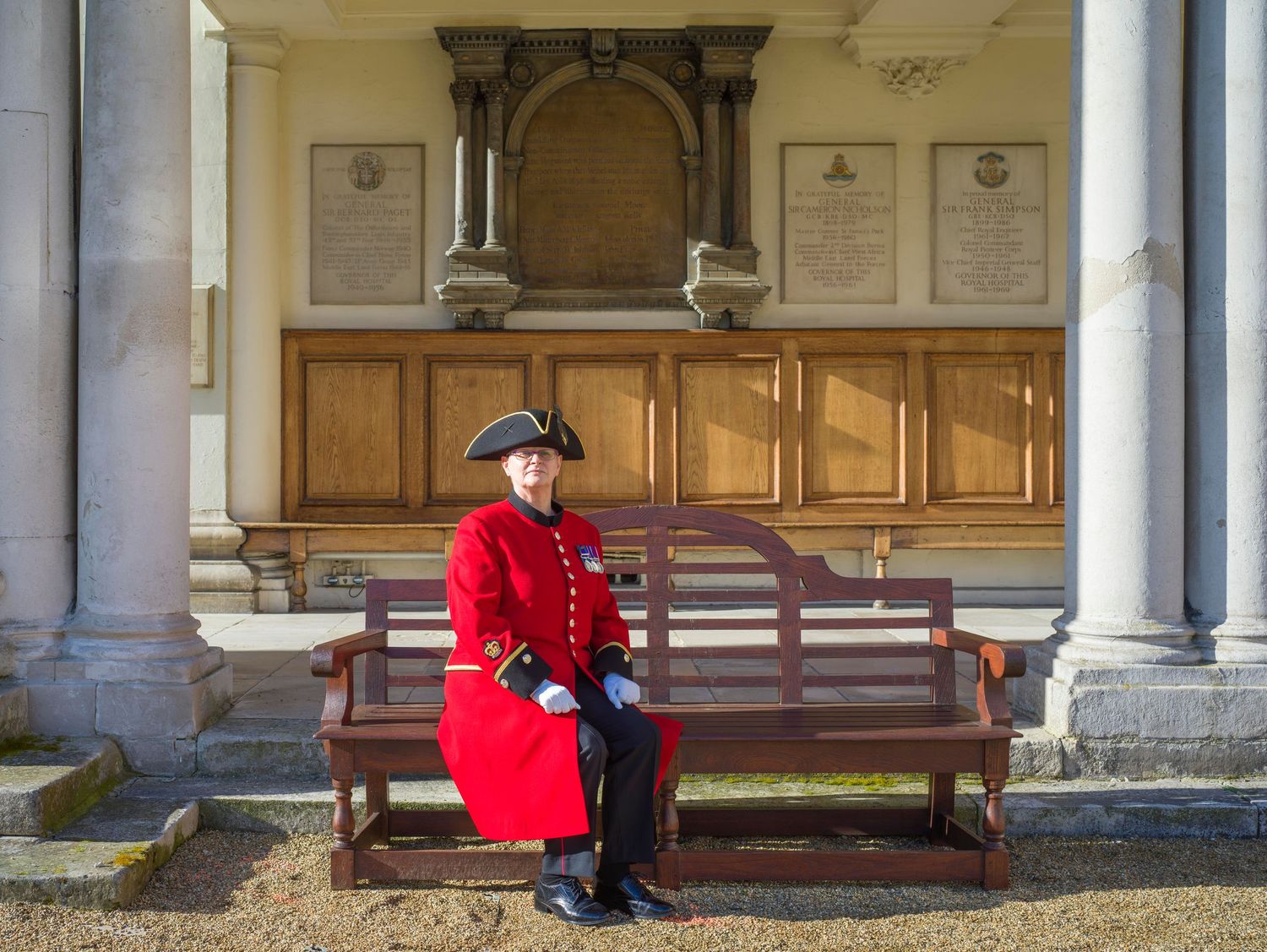 A Chelsea Pensioner in traditional red uniform sits on a wooden bench outside a historic building.