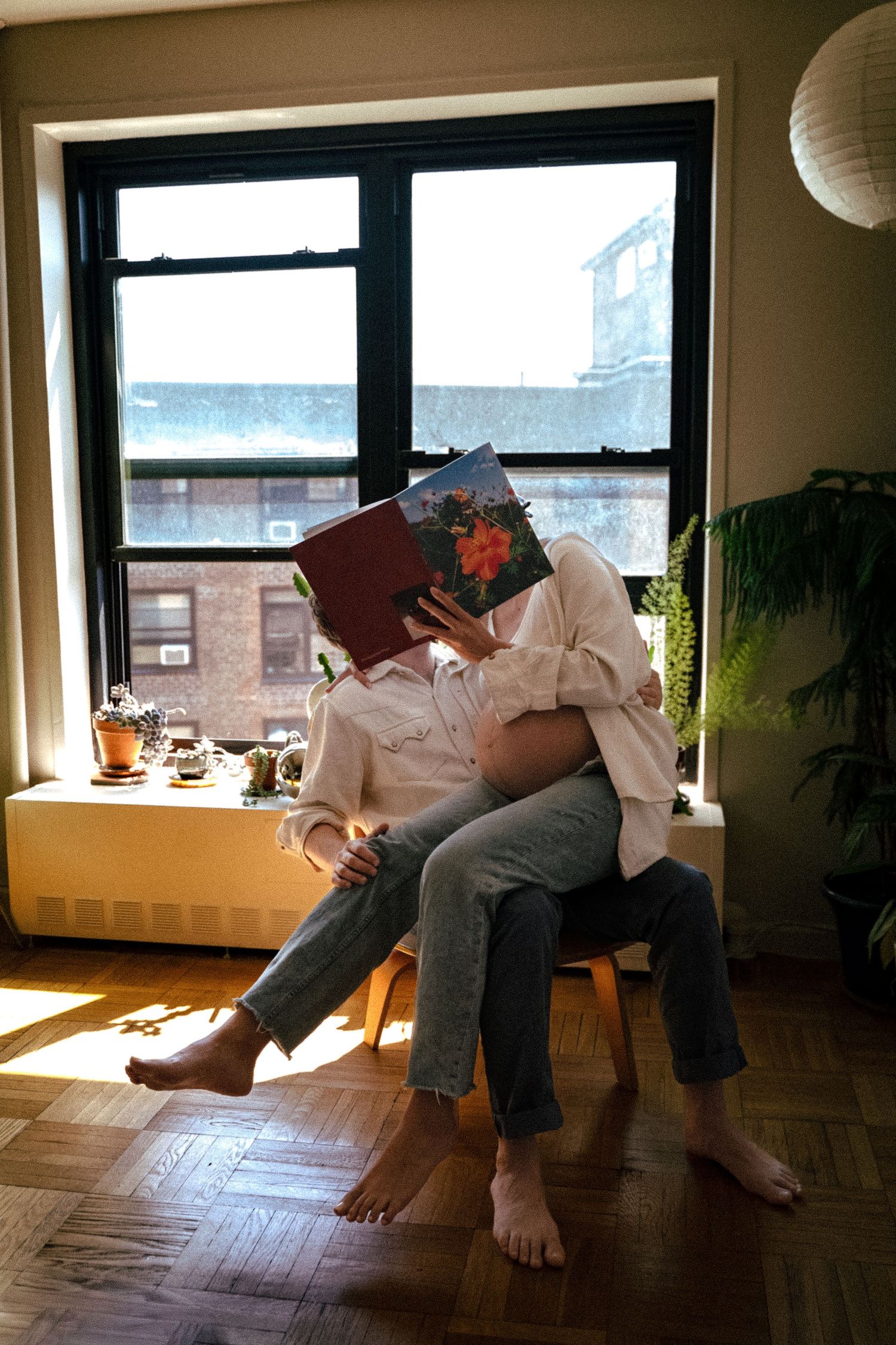 Someone relaxes barefoot in a sunny apartment room while reading a book near a window.