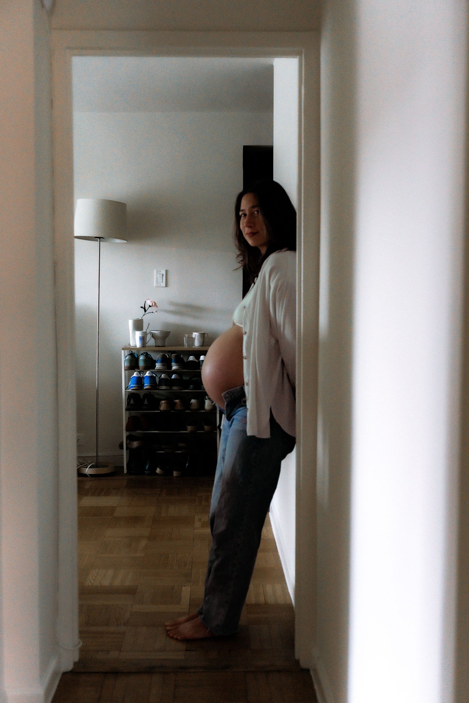 A pregnant woman in white stands in a hallway against a wall beside a floor lamp and shelving unit.