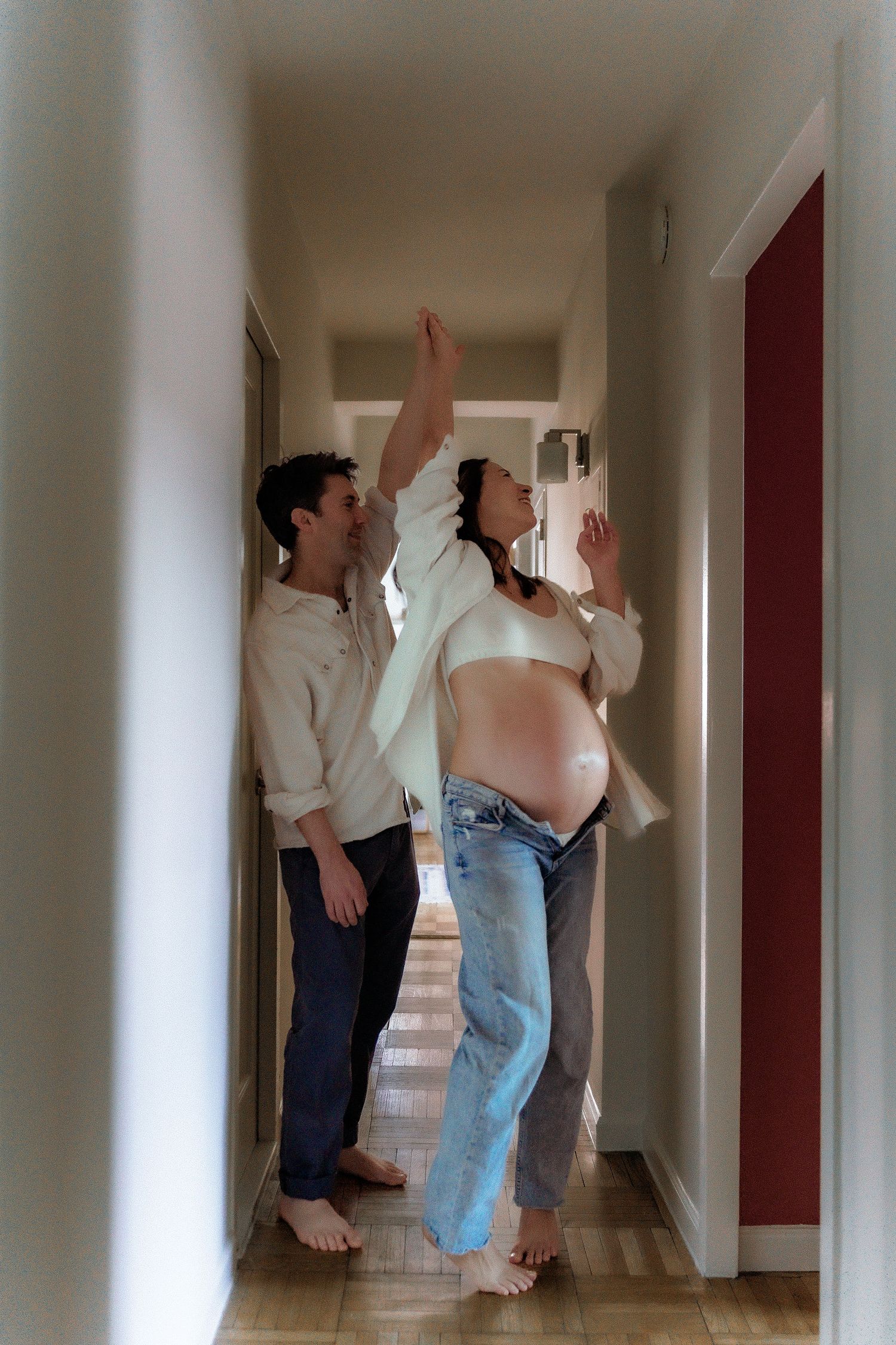 A couple stands together in a hallway, with the person on the right displaying a pregnant belly and reaching upward.