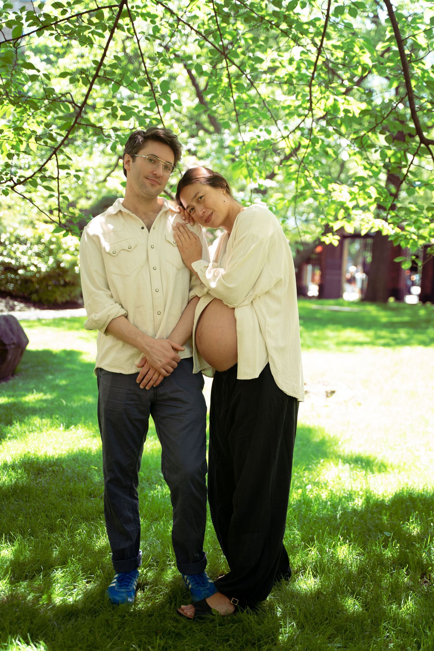 A couple stands together under green trees on a sunny spring day in a garden setting.