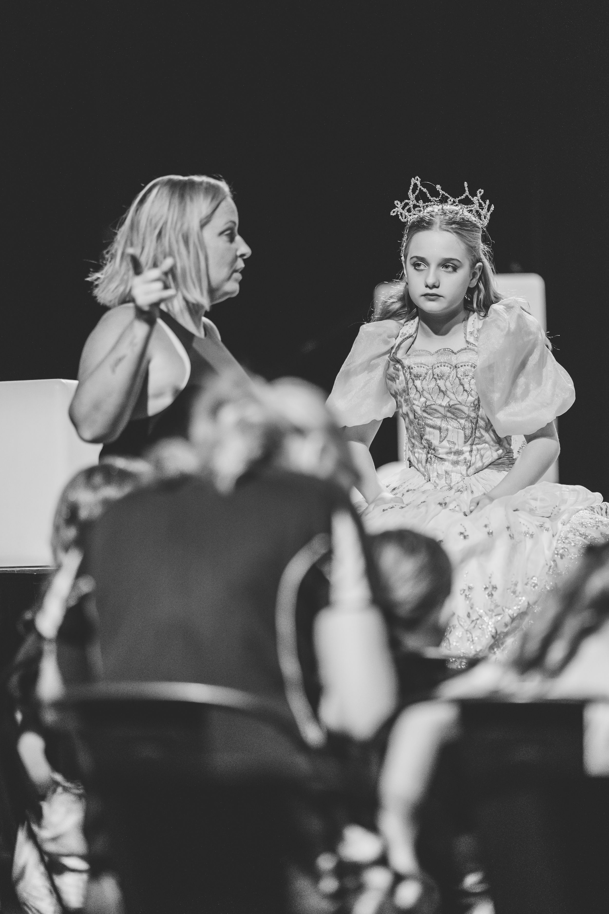 A black and white photo of a theatrical performance showing a performer in a white dress with a crown and another in discussion.