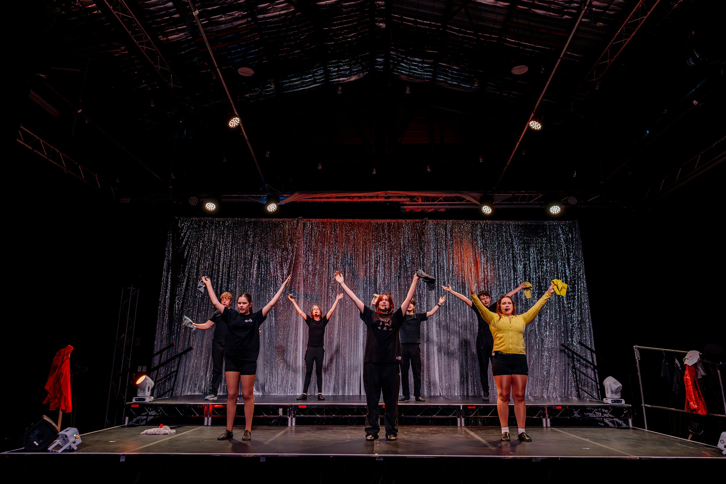 Performers in yellow and black outfits take a bow on stage in front of a silver curtain backdrop.