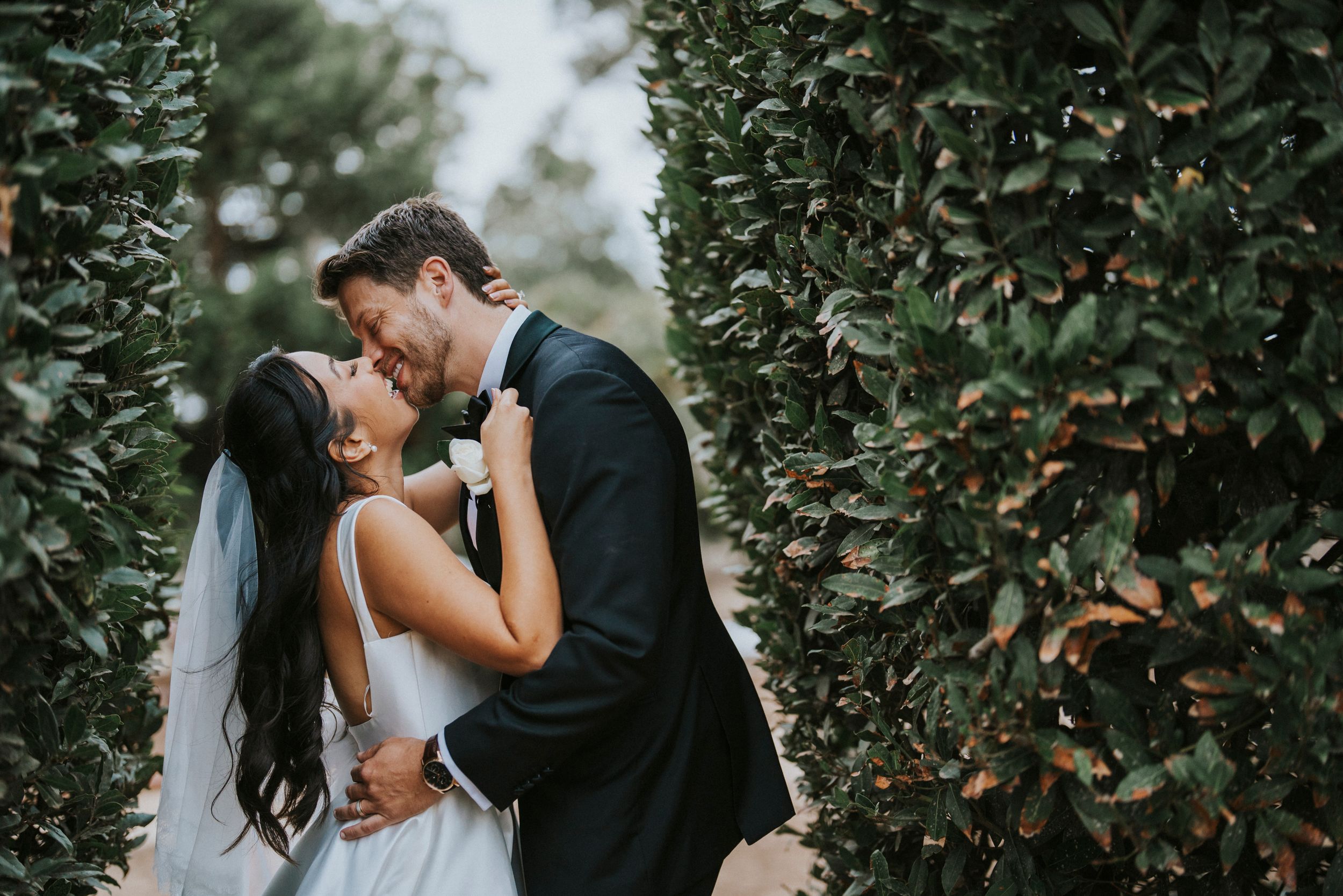 Newlywed couple sharing a romantic kiss among lush green foliage in an outdoor garden setting.