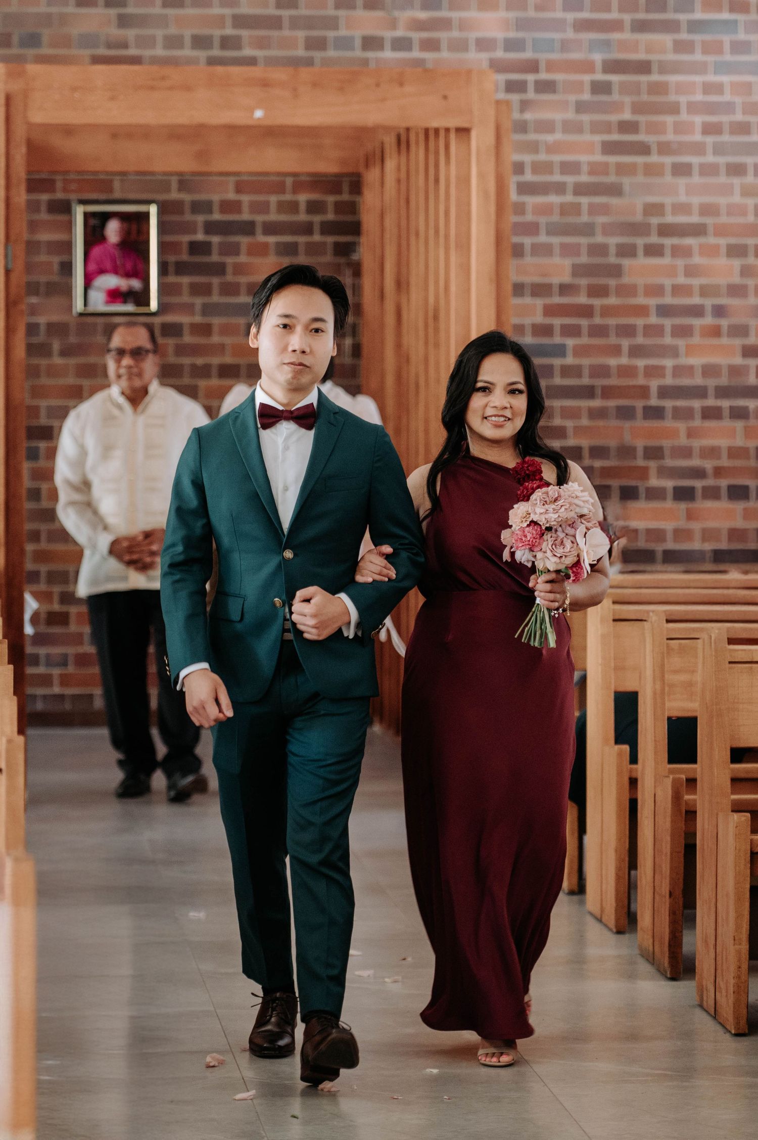 Wedding party members walk down church aisle with bride wearing burgundy dress and groom in emerald green suit.