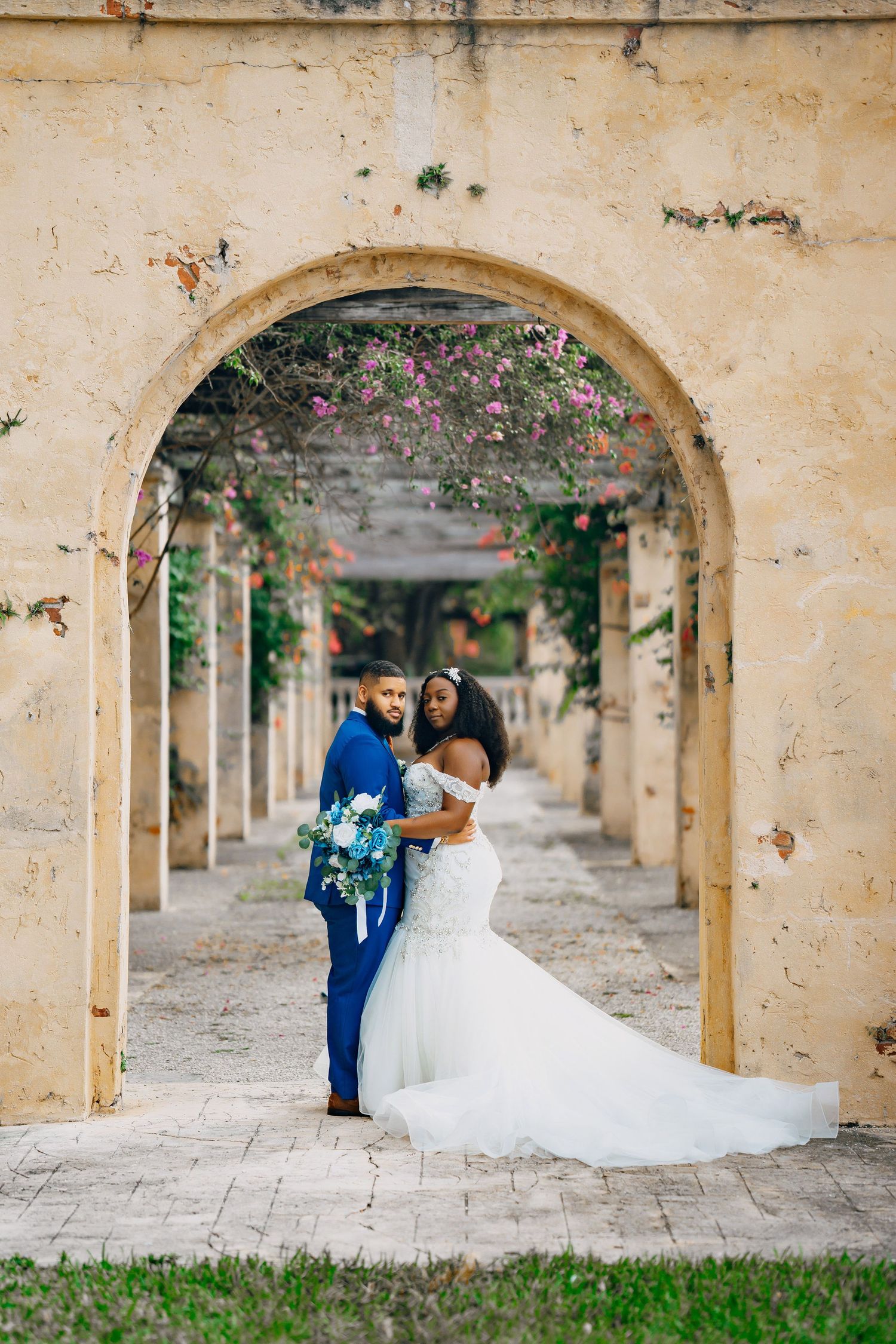 A romantic wedding moment captured under a stone archway with cascading flowers and Mediterranean architecture.