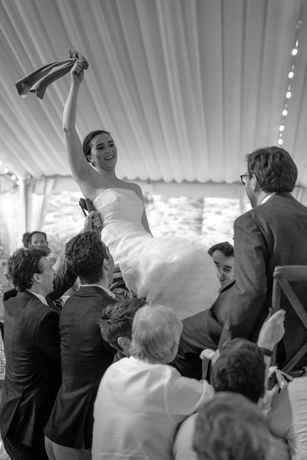 Bride lifted by wedding guests during celebratory hora dance inside white tent reception.