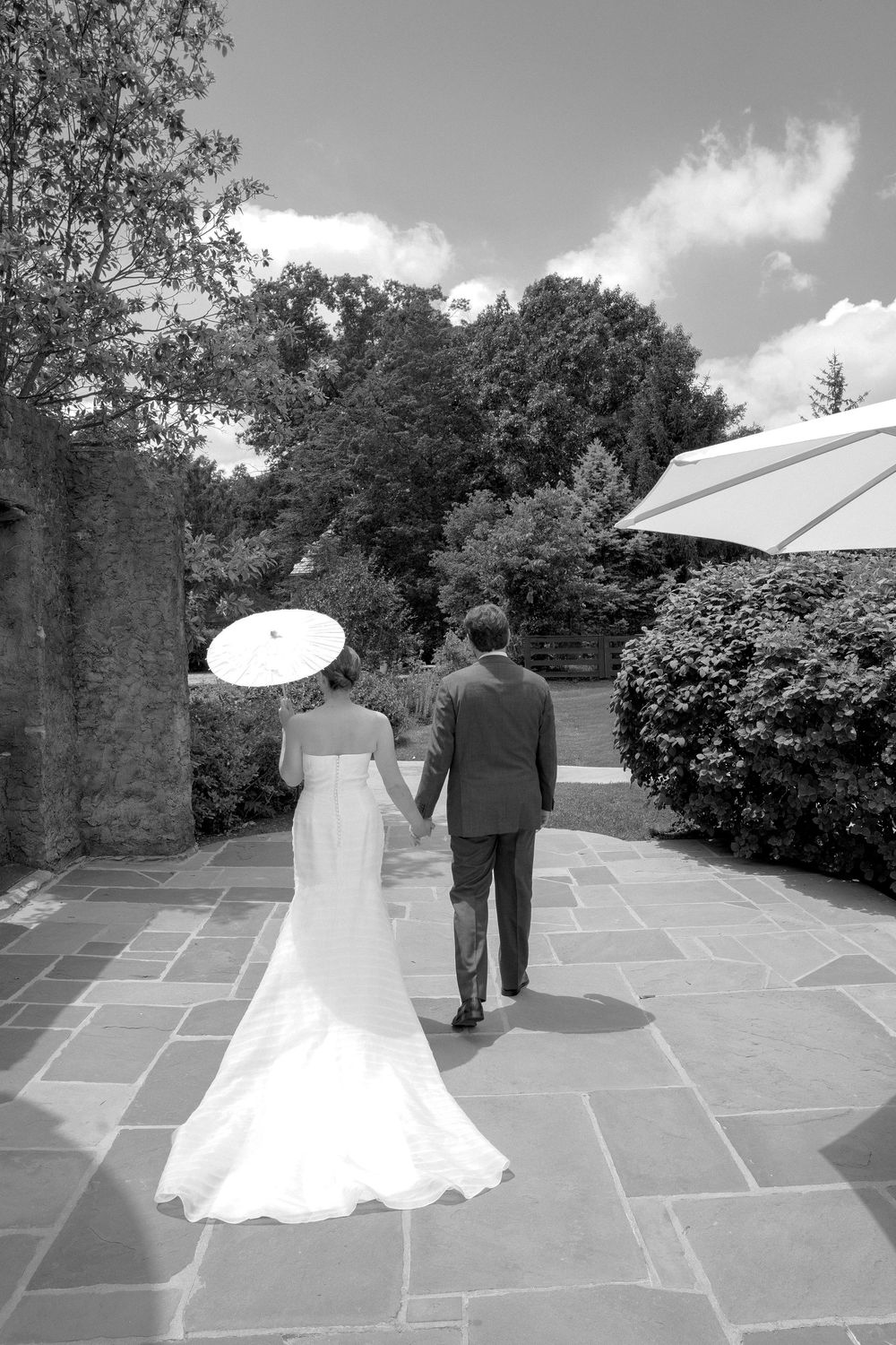 Bride and groom walk hand in hand on stone patio with parasol on sunny wedding day.