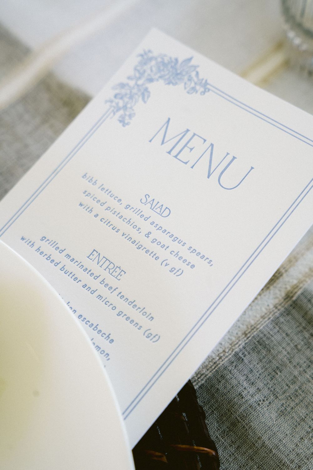 Elegant wedding menu card in blue and white featuring salad and entree options displayed on a linen tablecloth.