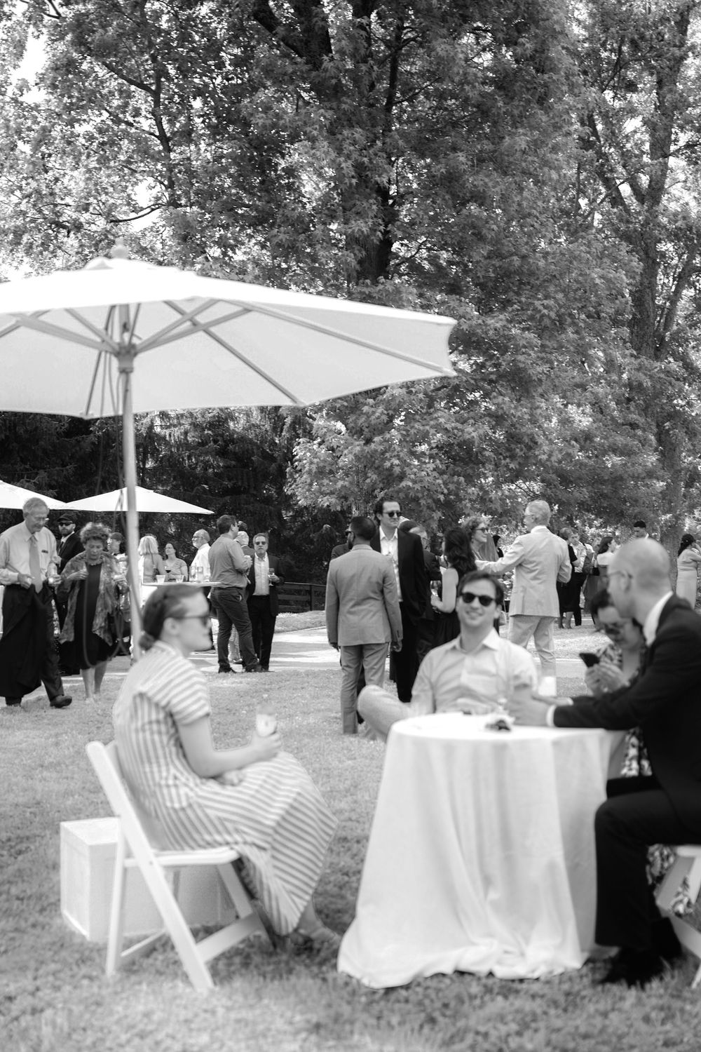 Black and white photo of wedding cocktail hour guests mingling outdoors under white umbrellas on lawn.
