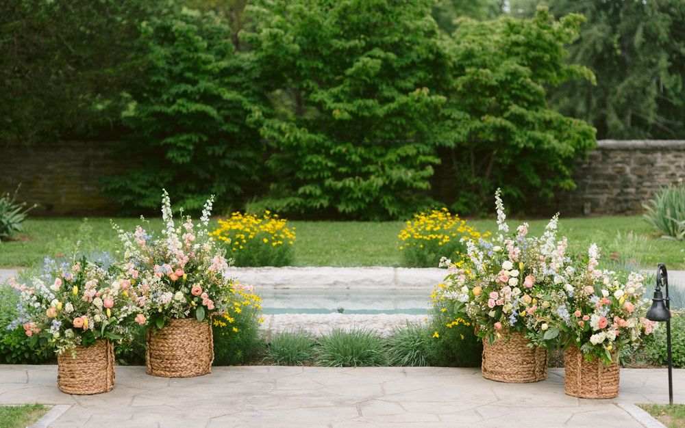 Four wicker baskets filled with lush pink, white, and yellow floral arrangements decorating an outdoor wedding ceremony.
