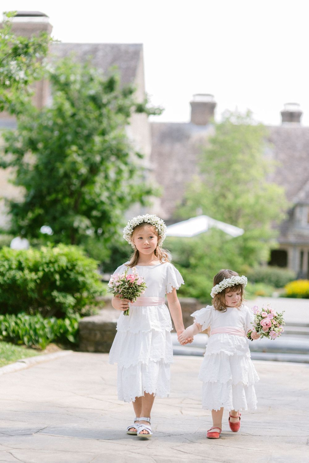 Two flower girls in white dresses and floral crowns walk hand-in-hand carrying pink bouquets outdoors.