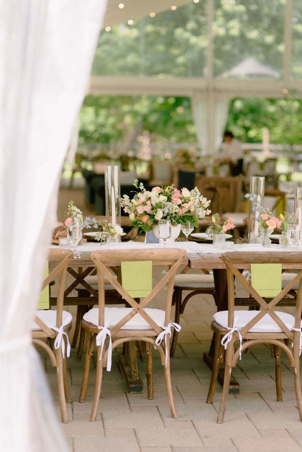 Rustic wooden farm tables with crossback chairs, white cushions, and pink floral centerpieces under an elegant outdoor wedding tent.