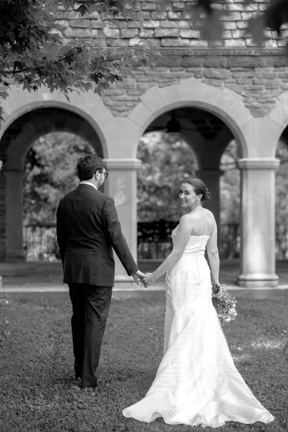 Bride and groom hold hands before stone arches in a classic black and white wedding portrait.