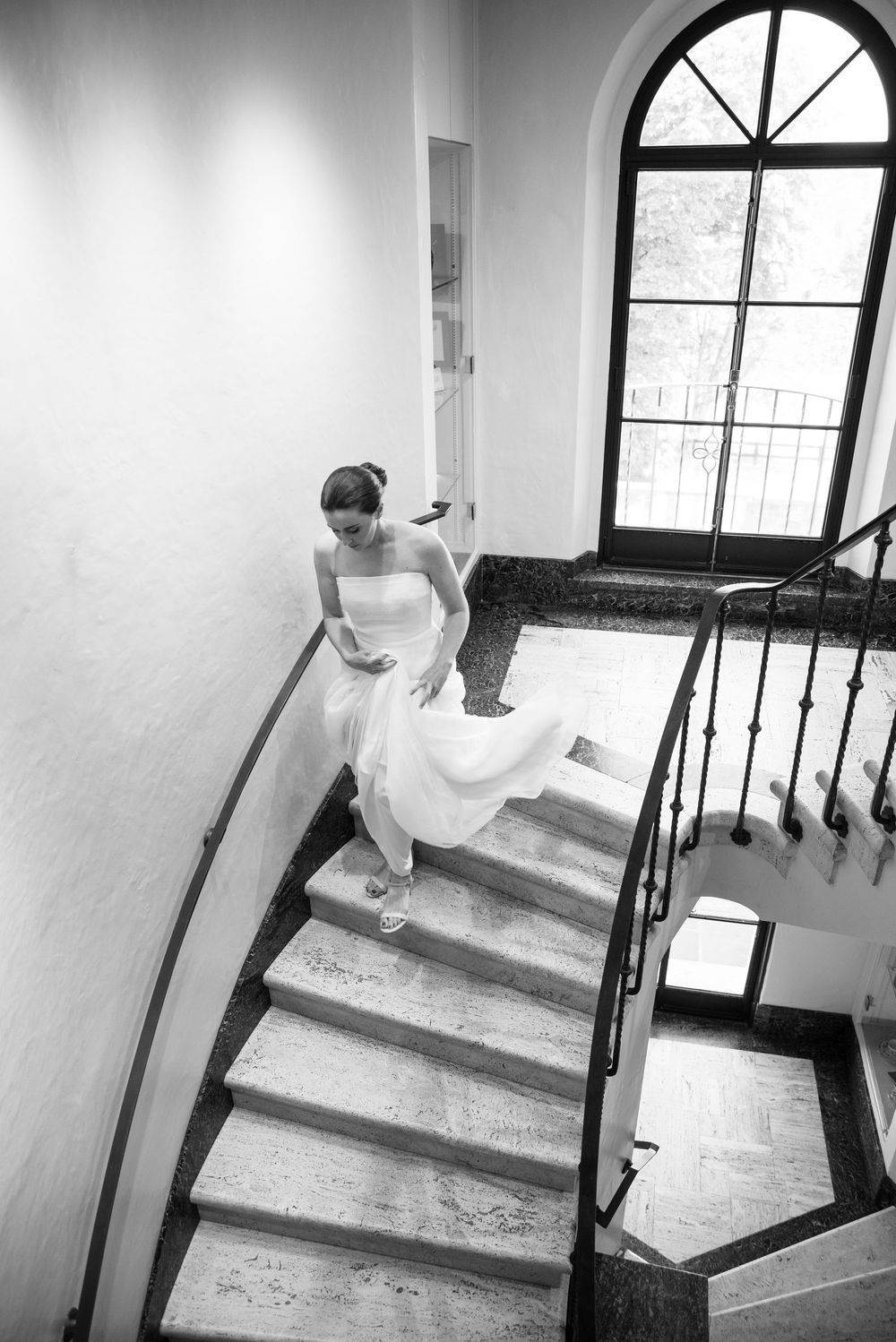 Bride in white gown descending a curved marble staircase with iron railing in a classic venue, black and white photo.