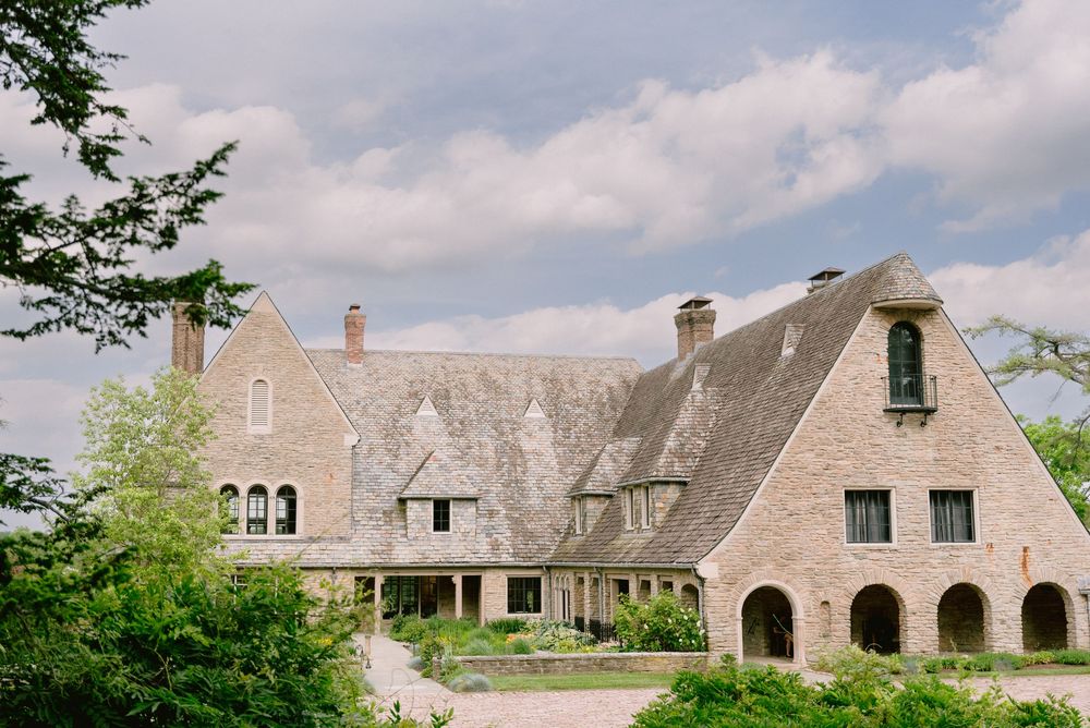 Historic stone manor house with steep gabled roof surrounded by mature trees under a partly cloudy sky.