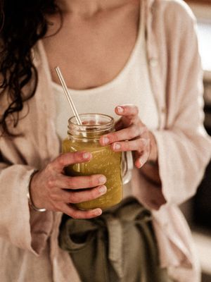 Person in cozy sweater holds mason jar of green smoothie with straw in warm lighting.