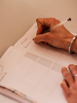 Someone wearing a bracelet writes in a planner or notebook with a pen, captured in a warm beige tone.