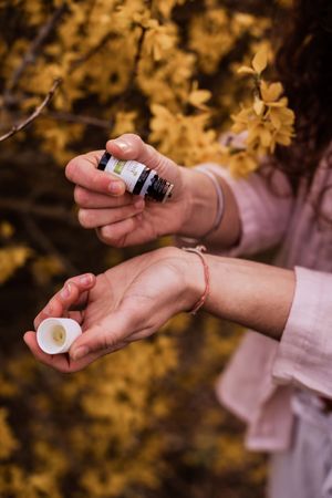 Hands pouring essential oil into palm wearing pink sleeves against autumn foliage background.
