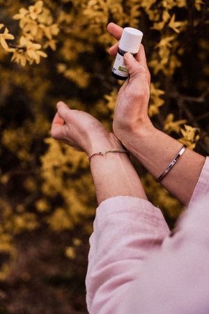 Hands holding small bottle against blurred golden autumn leaves and wearing delicate bracelets.