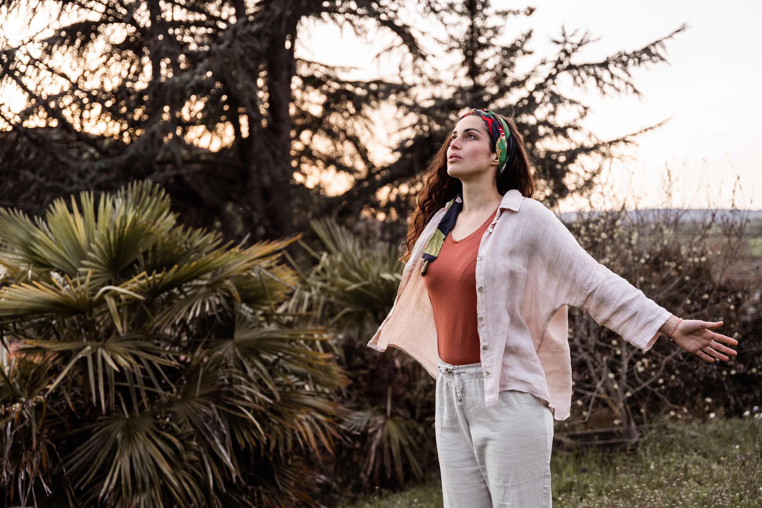 Person in white outfit enjoying nature with arms outstretched among palm trees at sunset.