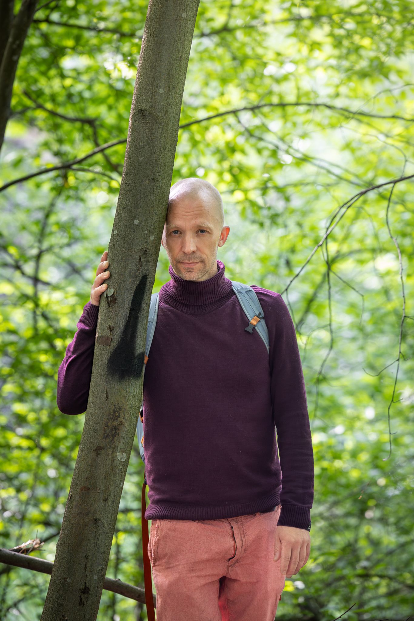 A person wearing a purple sweater and pink pants stands near a tree trunk in a lush green forest setting.