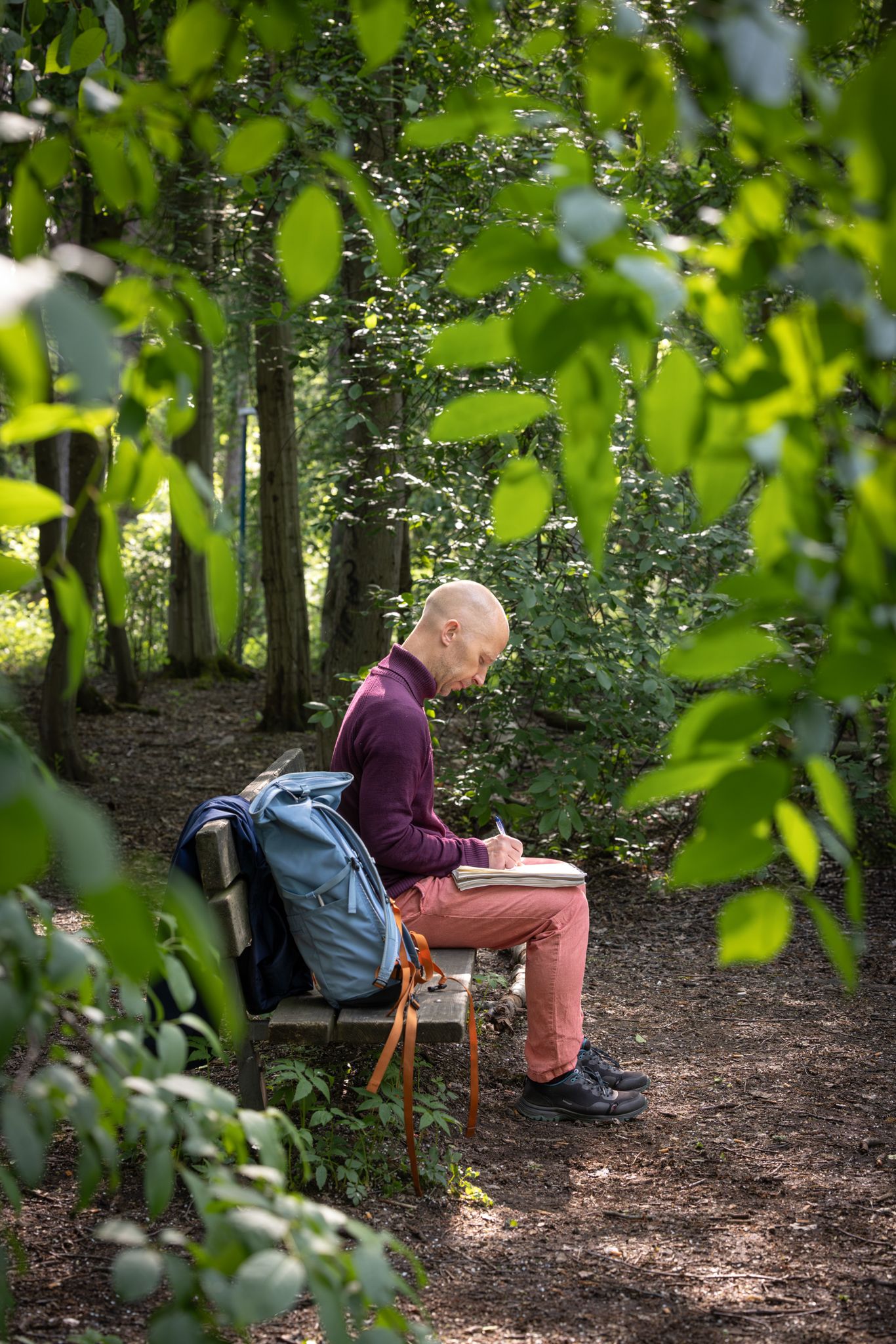 Person in purple shirt sits on wooden bench along forest trail with blue backpack and surrounded by green foliage.