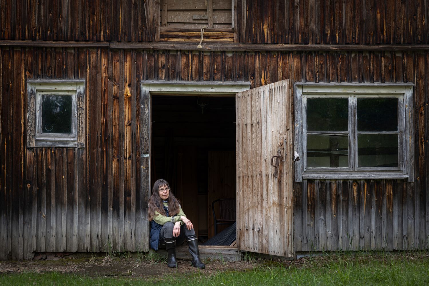 Person sitting in doorway of rustic weathered wooden barn with vintage windows in rural setting.