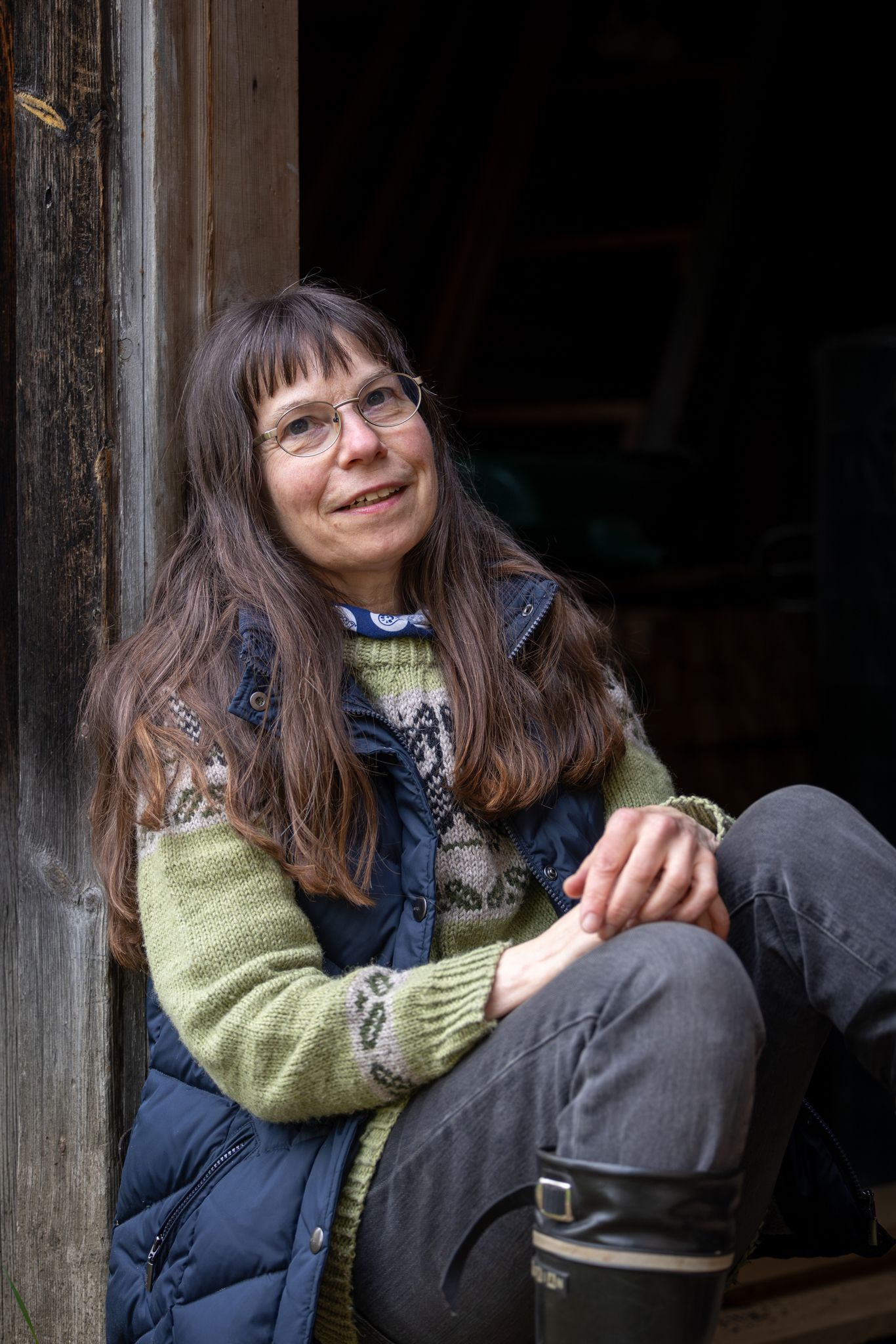 Woman with glasses and long brown hair smiling, seated in a barn doorway wearing a green sweater and vest.
