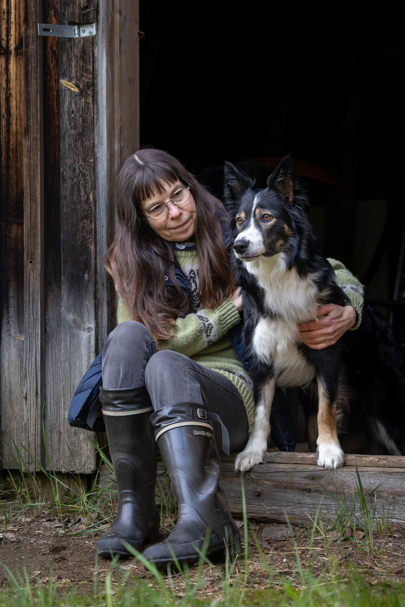 Woman in rubber boots sits with a tricolor Border Collie dog outside a rustic wooden barn door.