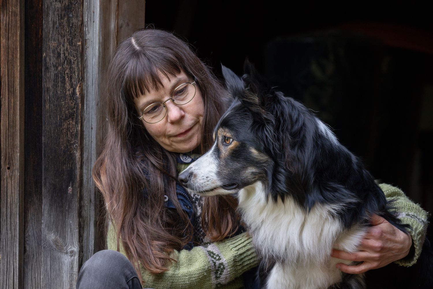 A woman with glasses tenderly embraces a black and white border collie dog near a rustic wooden structure.