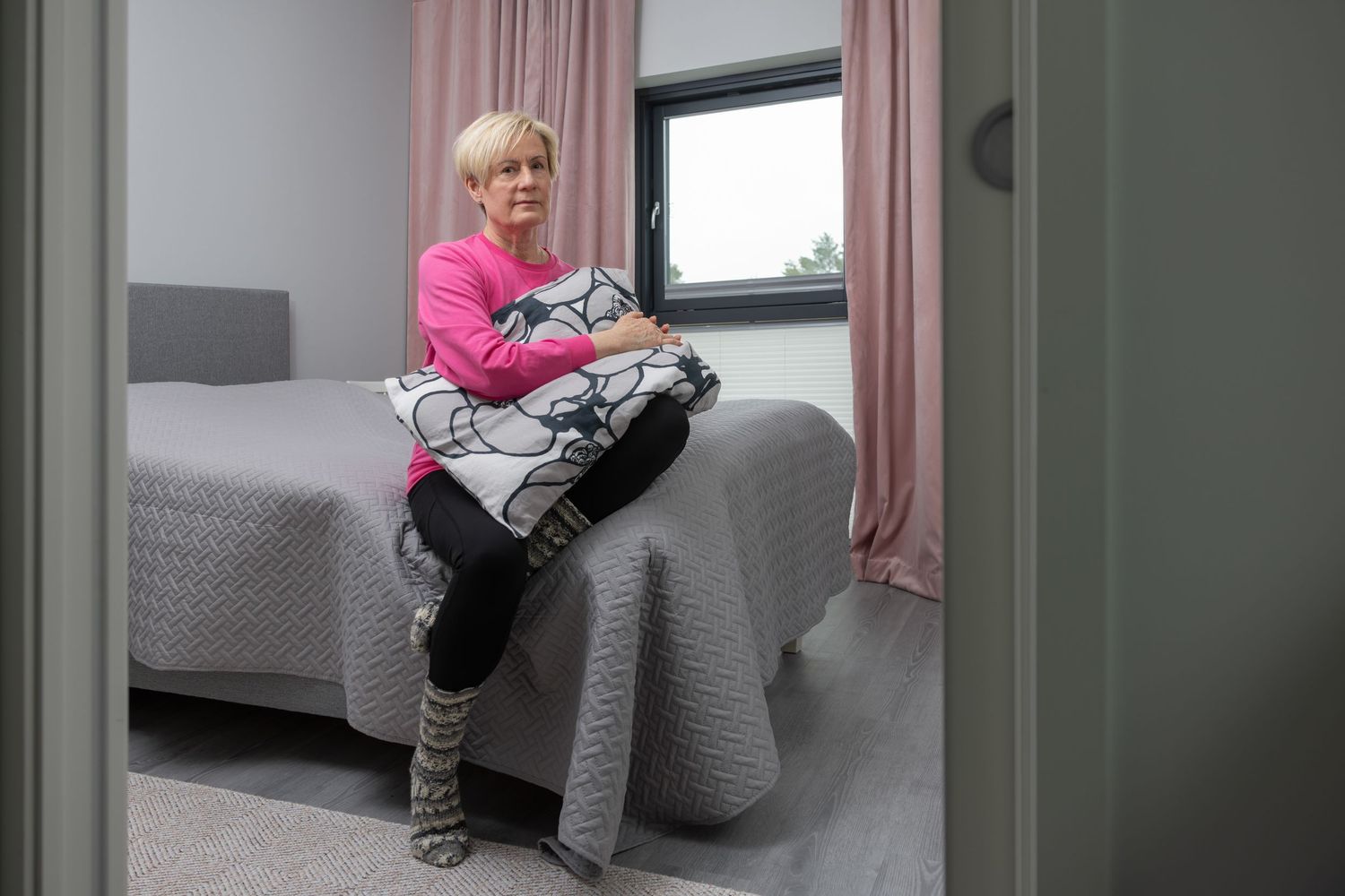 Woman in pink top sitting on bed holding decorative pillow in modern gray bedroom with curtains.