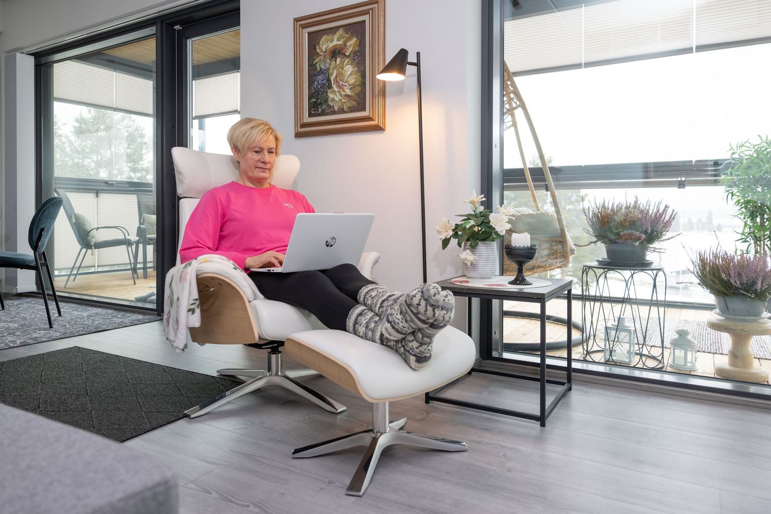 Woman in pink sweater using laptop in modern white recliner chair with footrest in bright living room.