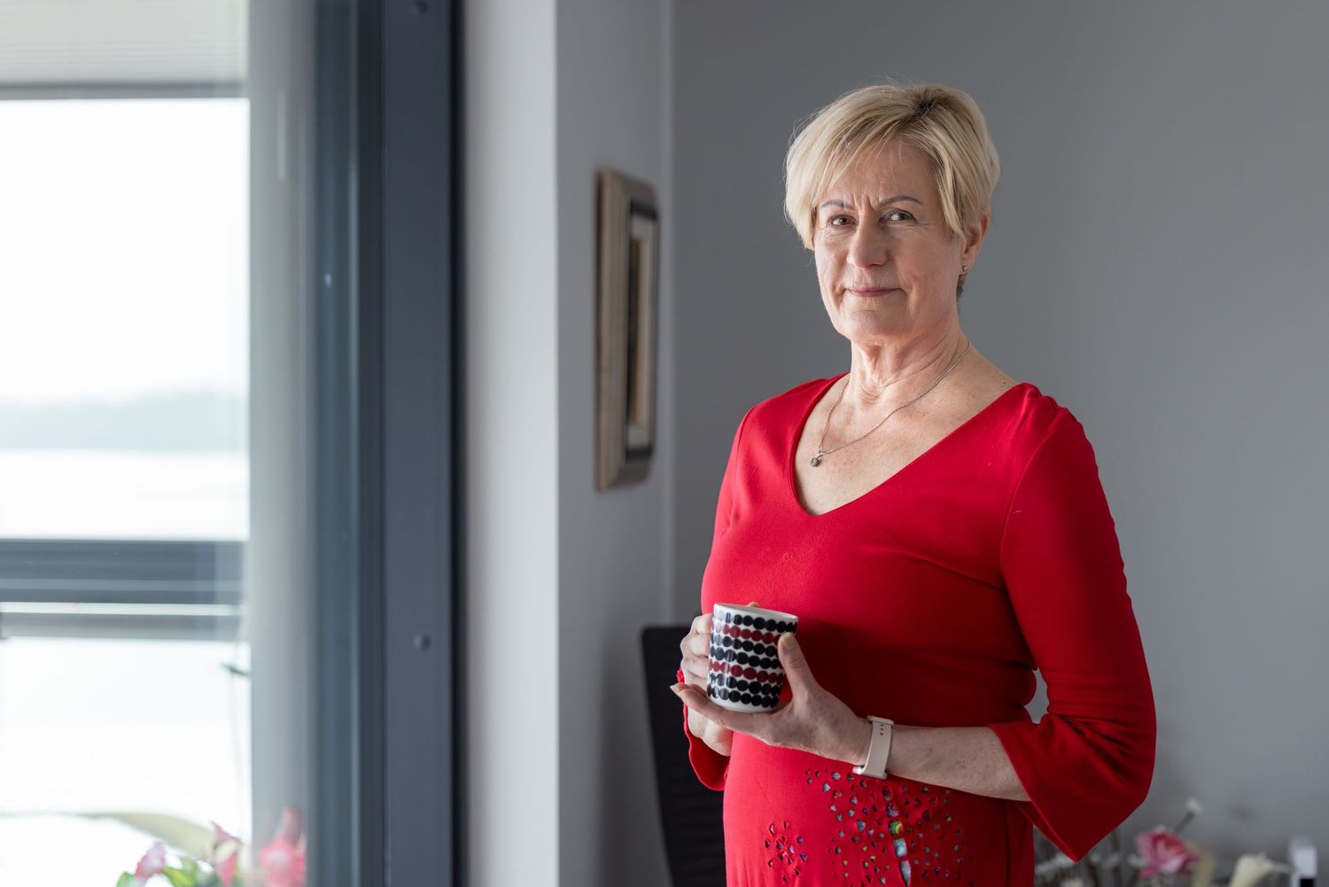 Middle-aged blonde woman in red dress holding coffee mug standing by window in modern home.